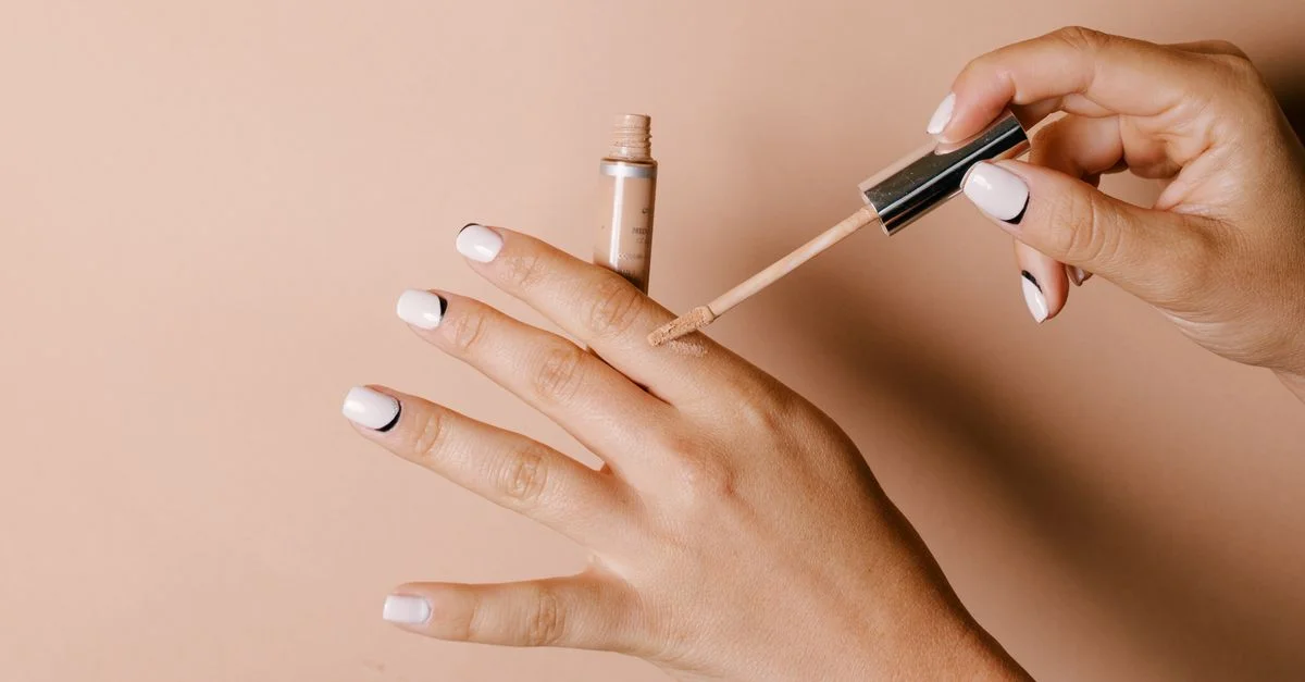 Des mains élégamment manucurées appliquent du correcteur liquide sur la peau sur un fond neutre.
