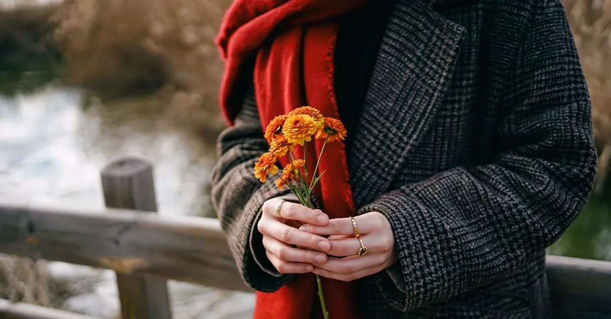 Une femme portant un manteau et une écharpe rouge tenant des fleurs orange sur un pont en bois à l'extérieur.